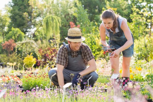 In a well-maintained garden in Ilford, two women are engaging in gardening activities amidst lush greenery and colourful flowering plants. The foreground features a flower bed filled with a variety of small, vibrant flowers, bordered by a neat edging. One woman, wearing a straw hat, a checkered shirt, and gardening gloves, is kneeling on the grass, tending to the flowers or plants. The other woman, dressed in a sleeveless top and denim overalls, and holding garden shears, is standing next to her, leaning slightly forward to assist or observe. Behind them, the garden showcases a mix of mature trees with leafy branches, well-trimmed hedges, and shrubs, creating a lush backdrop. The garden surface includes a grassy lawn with dense, green grass, and a stone or paved pathway runs slightly to the side, leading through the space. Bright, natural daylight illuminates the scene, indicating a clear, sunny day and contributing to a warm, inviting outdoor environment. This image reflects typical garden maintenance work, possibly undertaken by professional gardening services in Ilford, with an emphasis on vibrant plant life, tidy landscaping, and outdoor enjoyment.