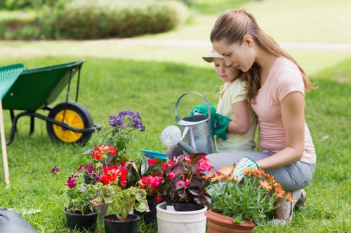 A woman and a young girl are kneeling on a well-maintained, lush green lawn in a garden, engaged in planting and tending to a variety of potted flowers and plants, which include red, purple, and orange blooms. The garden features a neatly trimmed hedge in the background, with a paved pathway visible beyond, and a green wheelbarrow nearby, indicating outdoor maintenance activities. The scene is set under natural daylight with a soft, clear sky, creating a bright and inviting outdoor environment typical of a residential garden in Ilford. This image emphasizes garden care, planting, and outdoor attention, aligning with gardening services in the Ilford area such as maintenance, clearance, and landscaping offered by Gardening Services Ilford.